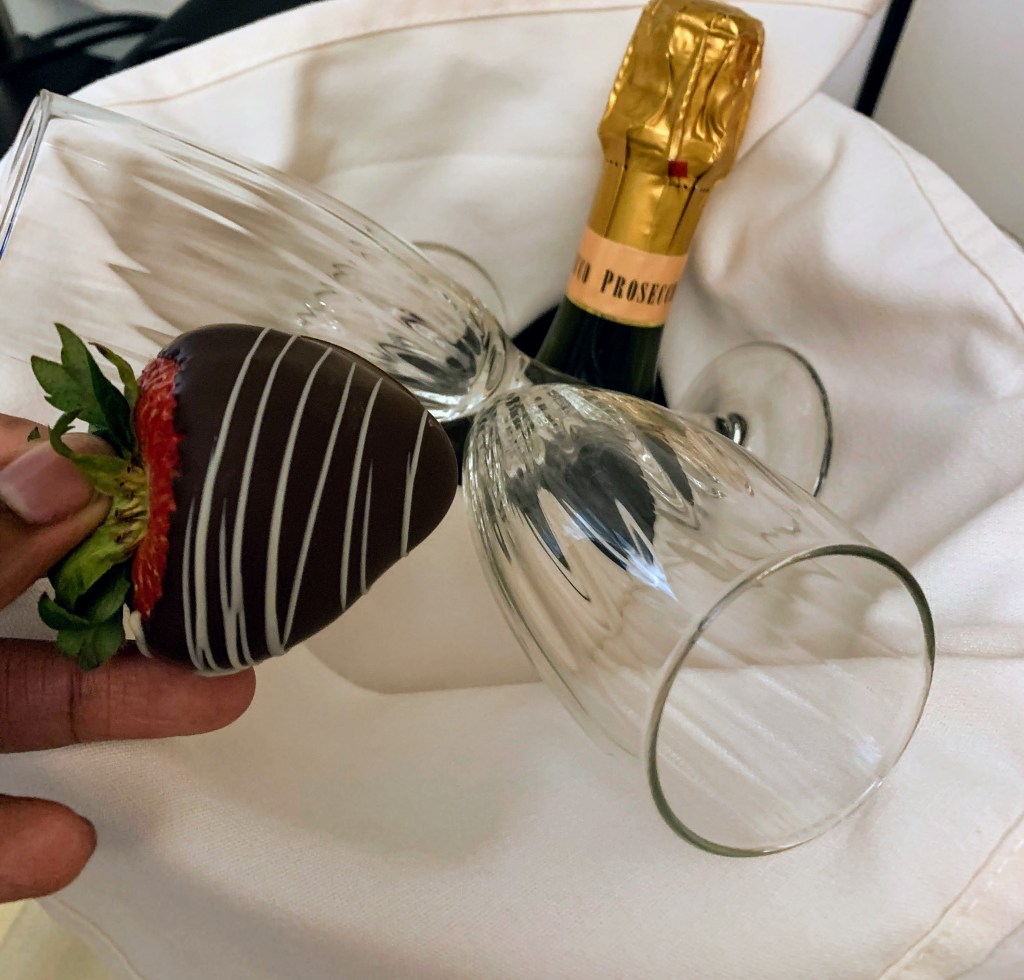 A chocolate covered strawberry, 2 glasses and a bottle of champagne on a white napkin background