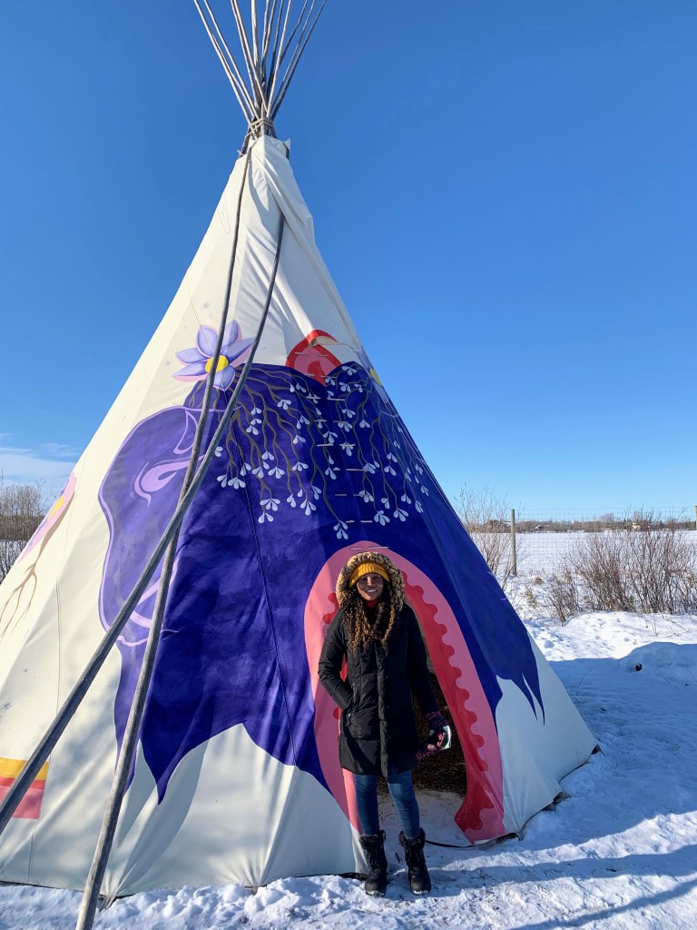 A person in a winter coat stands in front of a traditional indigenous shelter.